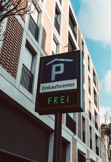 A vertical parking sign mounted on a pole in front of a modern multi-storey building during daytime. The sign displays a large white letter 'P' with a blue background, indicating parking availability,