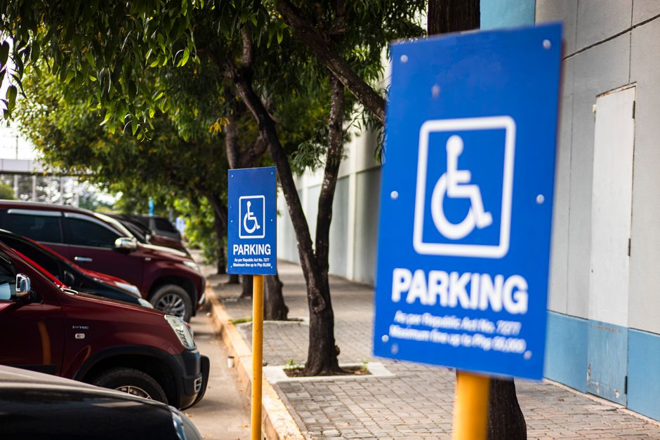 A vertical parking sign mounted on a pole in front of a modern multi-storey building during daytime. The sign displays a large white letter 'P' with a blue background, indicating parking availability,